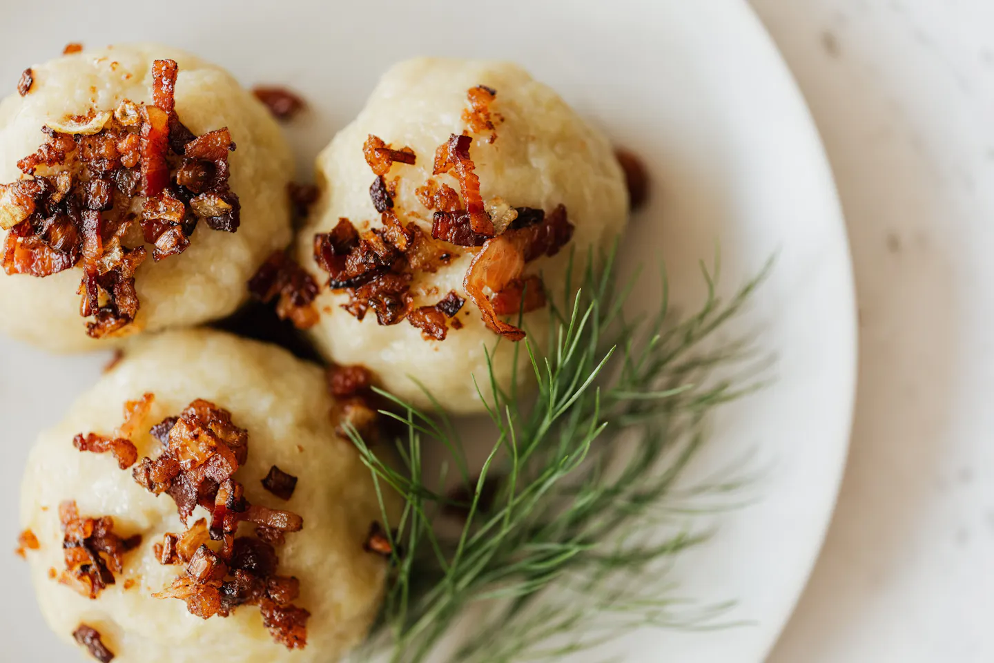 Three cepelinai potato dumplings topped with fried bacon and onion on a white plate beside a sprig of dill