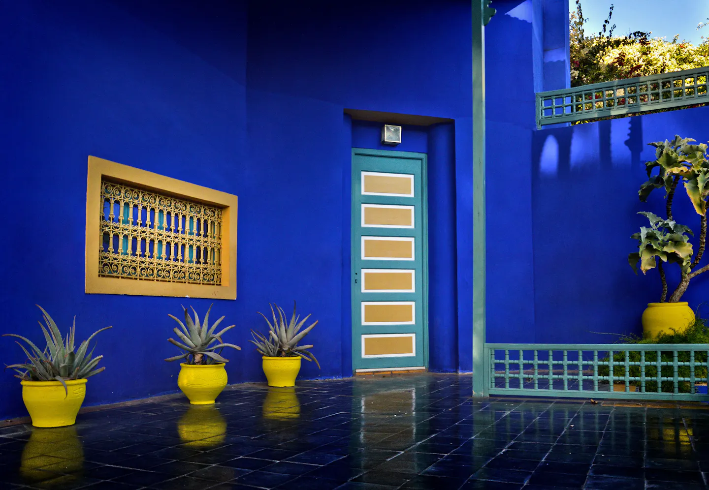 A Moroccan courtyard painted in vivid cobalt blue, with a turquoise door, a yellow-framed lattice window and aloe plants in bright yellow pots