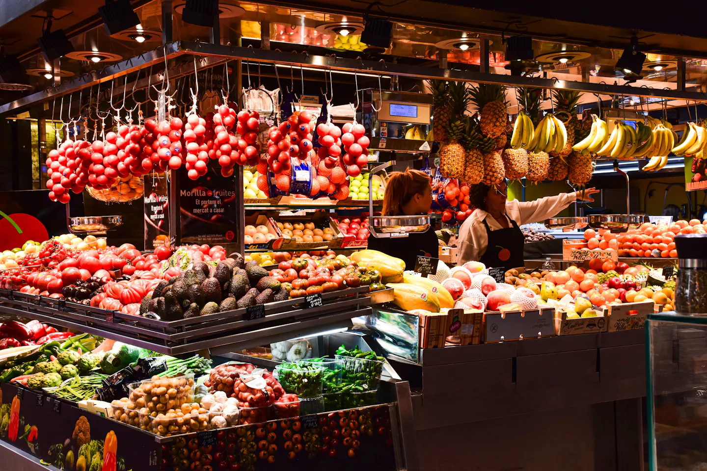 A fruit and vegetable stall in Barcelona's Boqueria market with hanging tomatoes, pineapples and bananas under warm lights