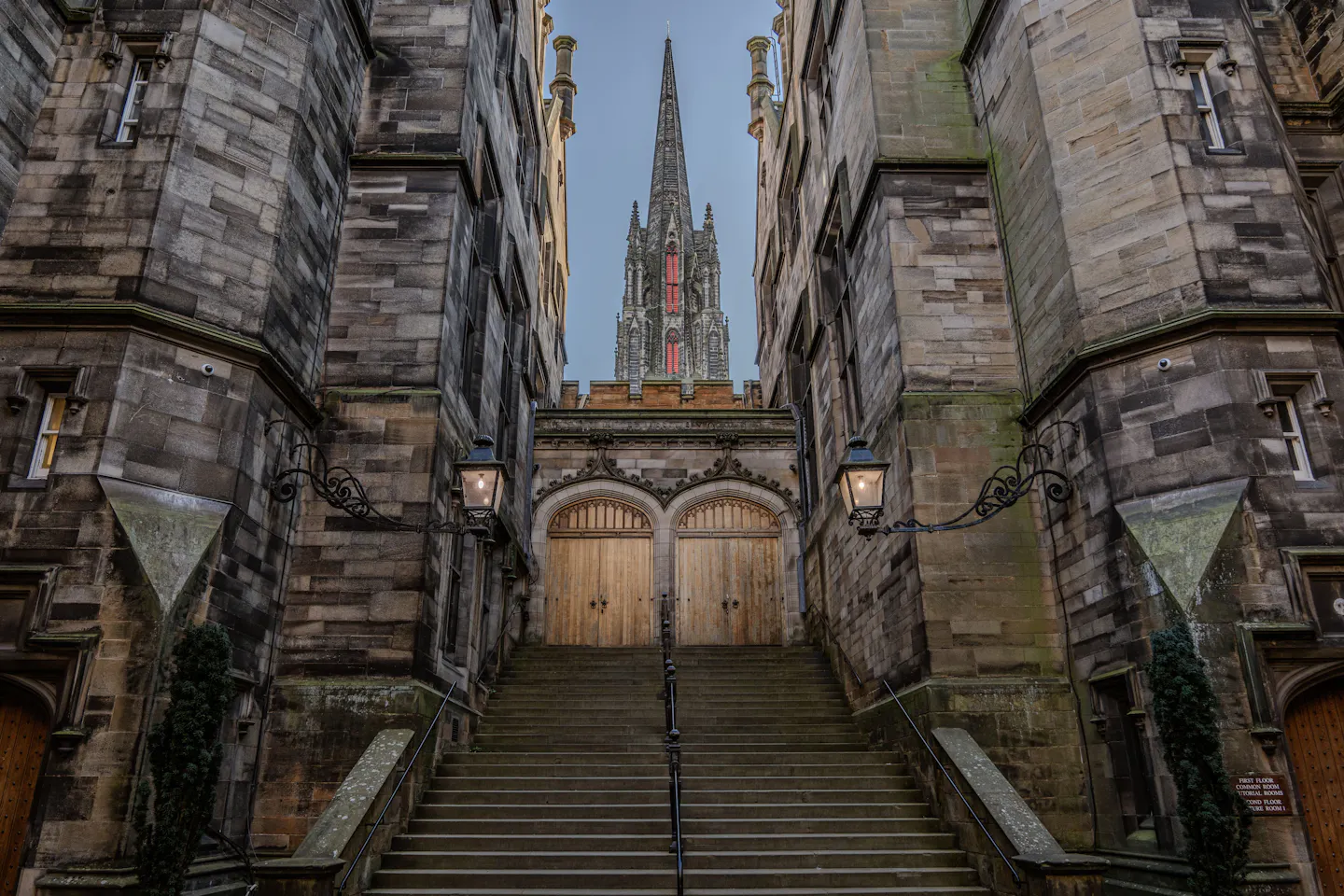 A steep flight of stone steps rising between dark tenement walls to twin lit lanterns and studded wooden doors, a gothic spire framed against blue dusk sky above