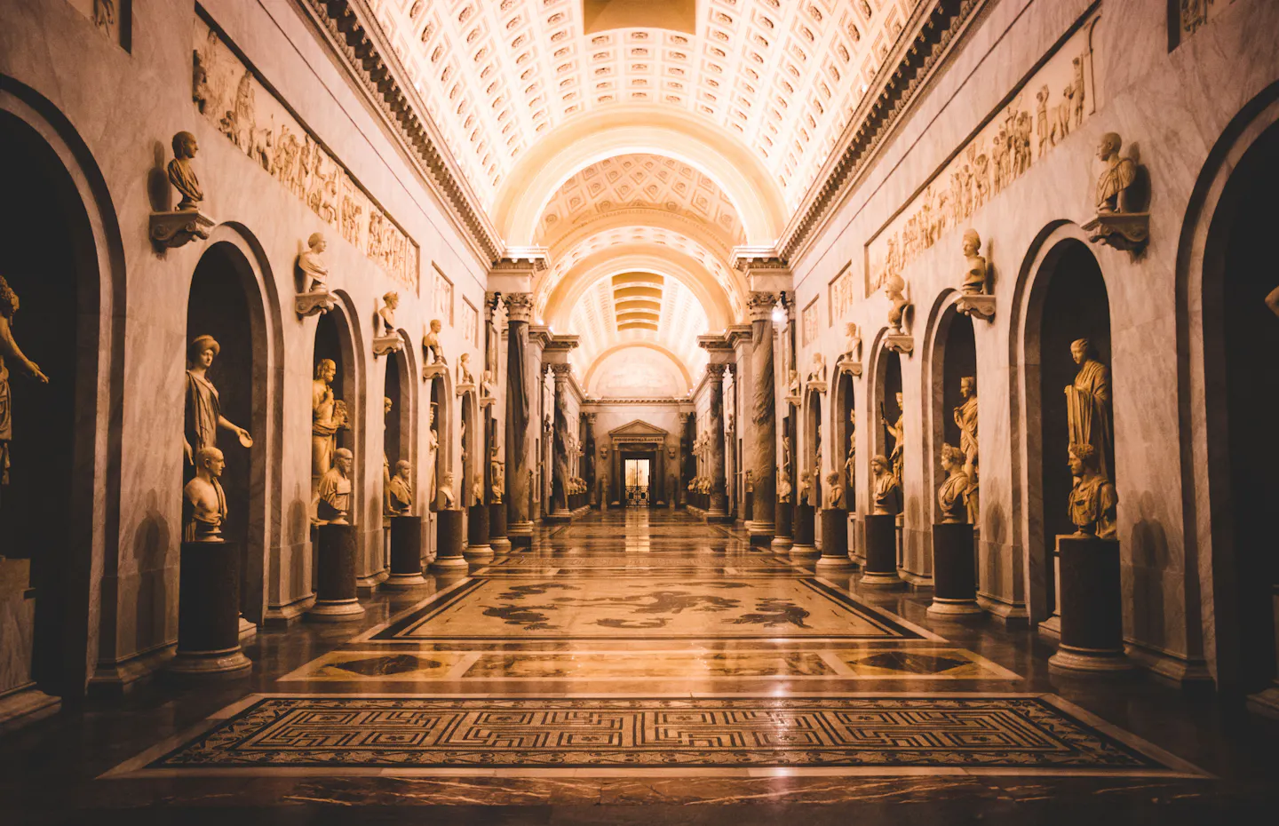 A long vaulted sculpture gallery in the Vatican Museums, with classical statues in niches along both walls and a mosaic marble floor