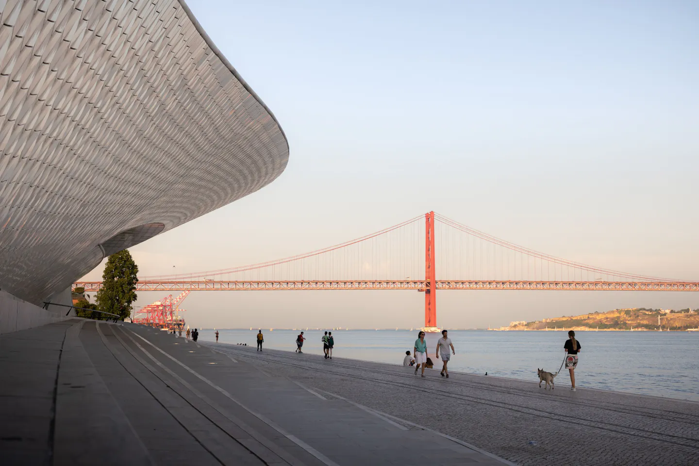 People strolling the Tagus riverfront in Lisbon at golden hour, one walking a small dog, with the red Ponte 25 de Abril behind