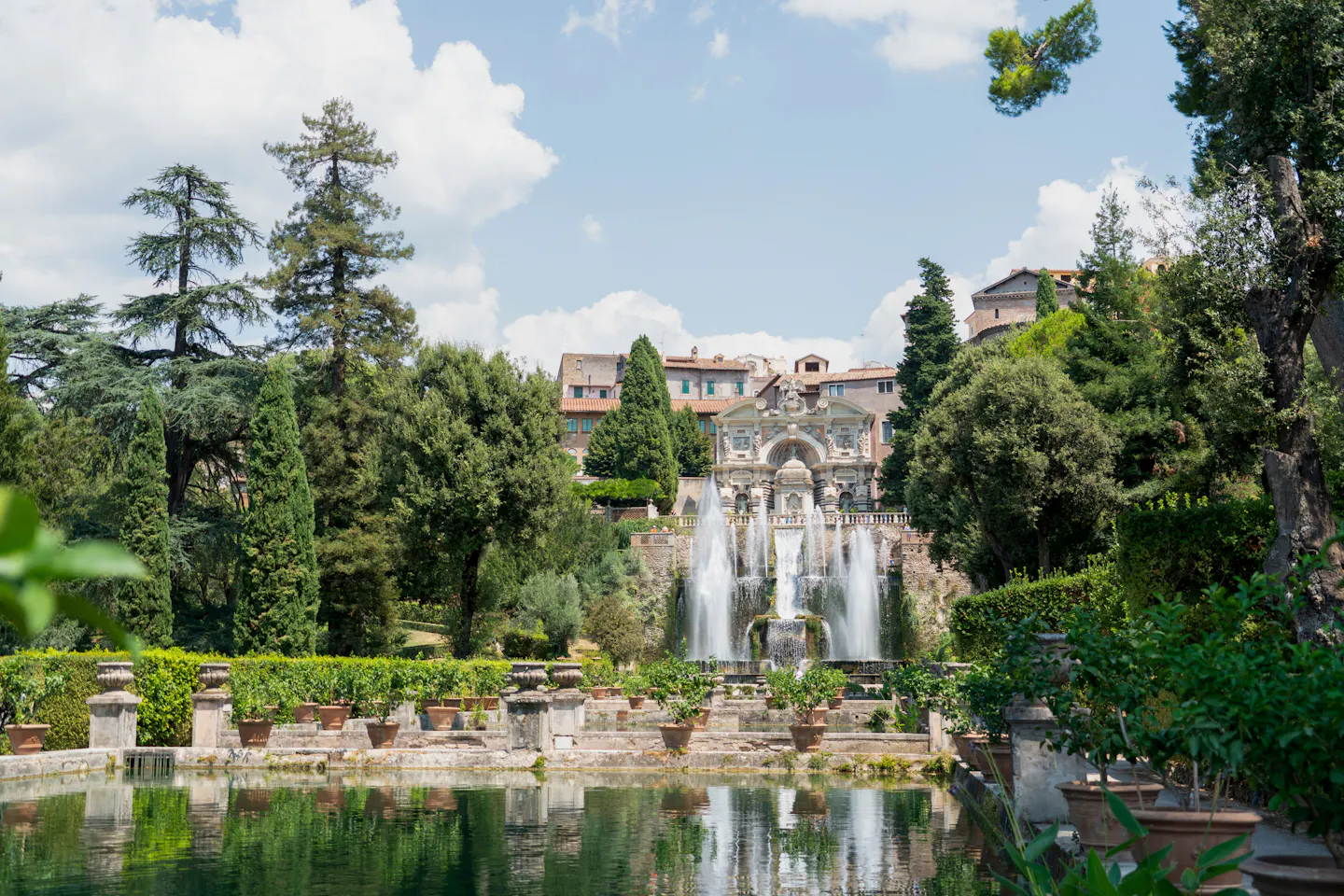 The Neptune Fountain at Villa d'Este in Tivoli throwing tall jets of water below the villa, framed by cypresses and cedars, with the still reflecting pool and rows of potted plants in the foreground