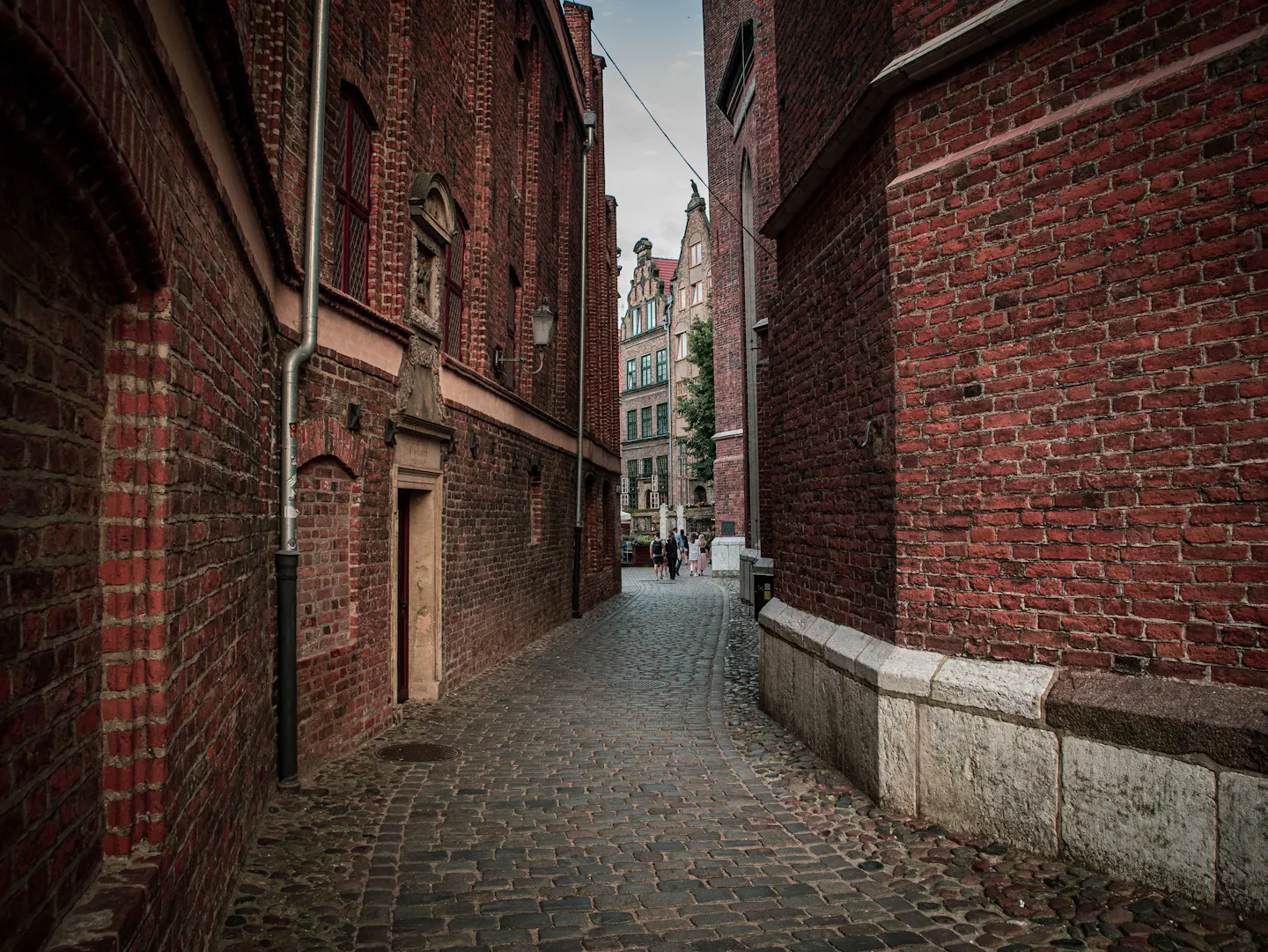 Narrow cobbled pedestrian lane lined with historic red-brick Gothic buildings, with a few walkers in the distance