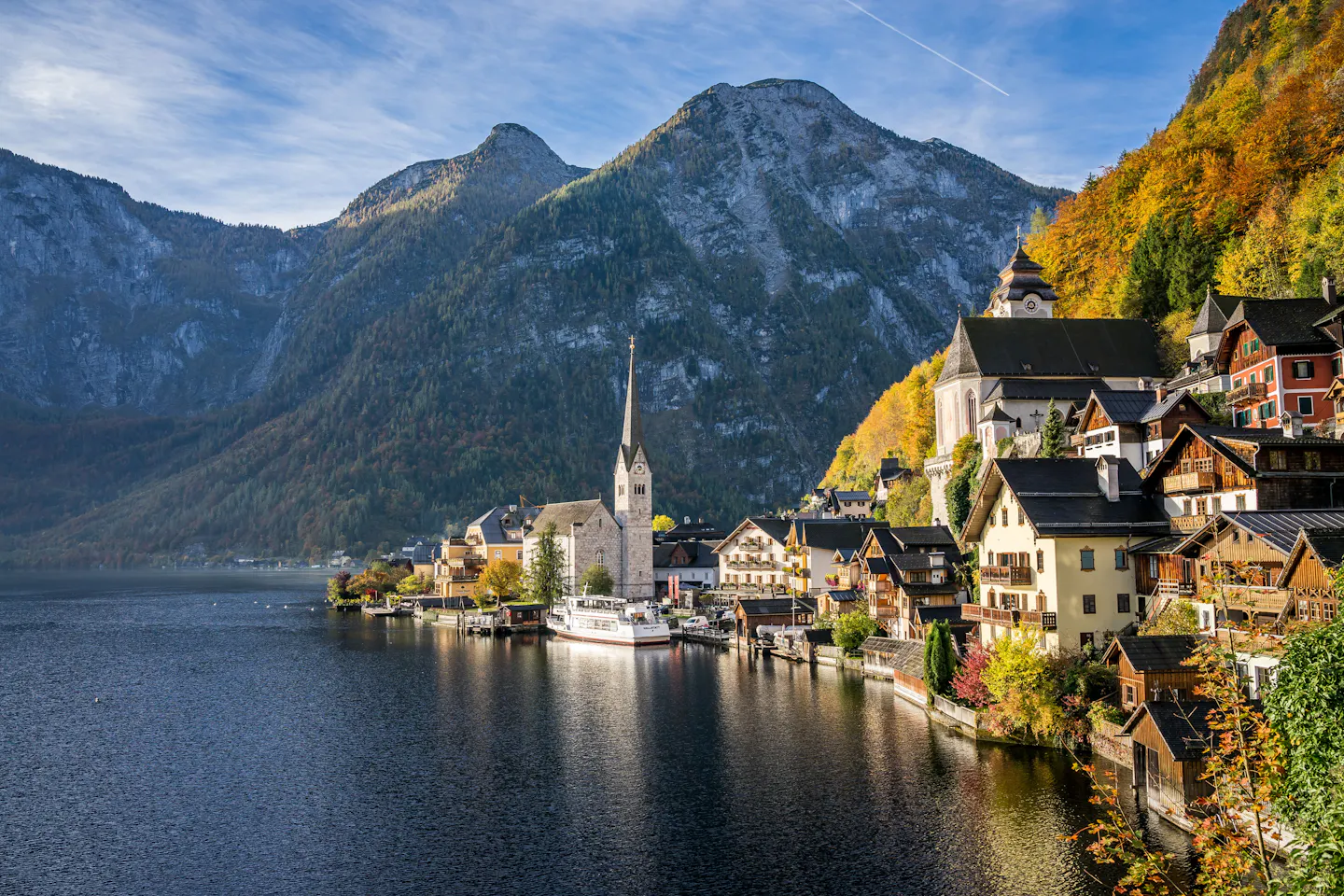 Hallstatt in autumn: wooden houses and the tall church spire stacked along the lakeshore, golden larches on the slope above and a grey mountain wall behind, reflected in the still Hallstätter See