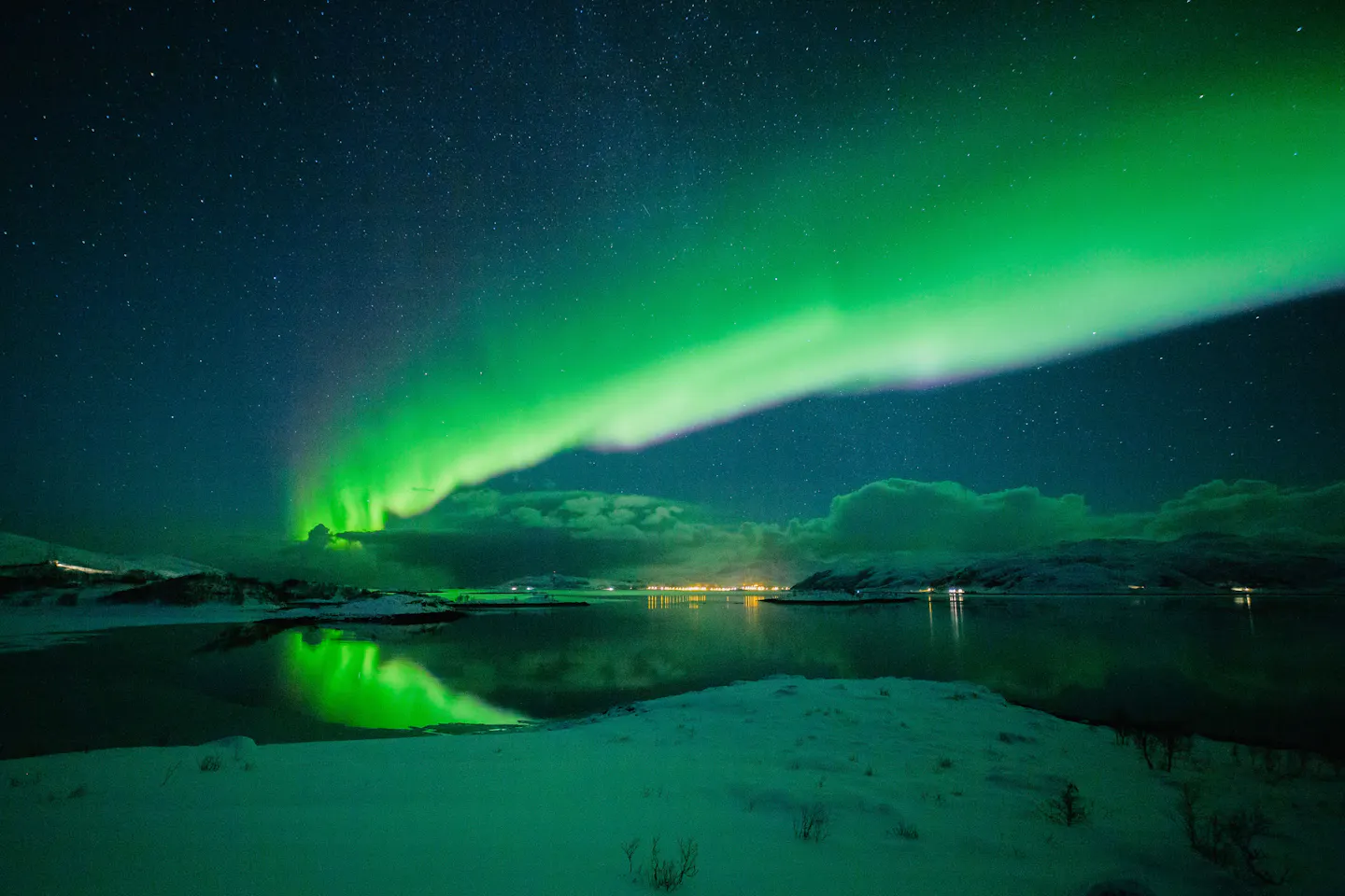 A wide green aurora arc over a snow-covered shoreline and dark fjord water near Tromsø in northern Norway