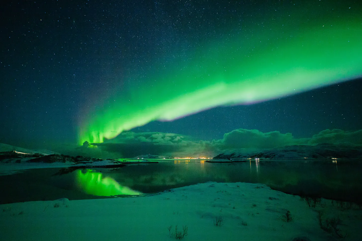 A wide green aurora arc over a snow-covered shoreline and dark fjord water near Tromsø in northern Norway