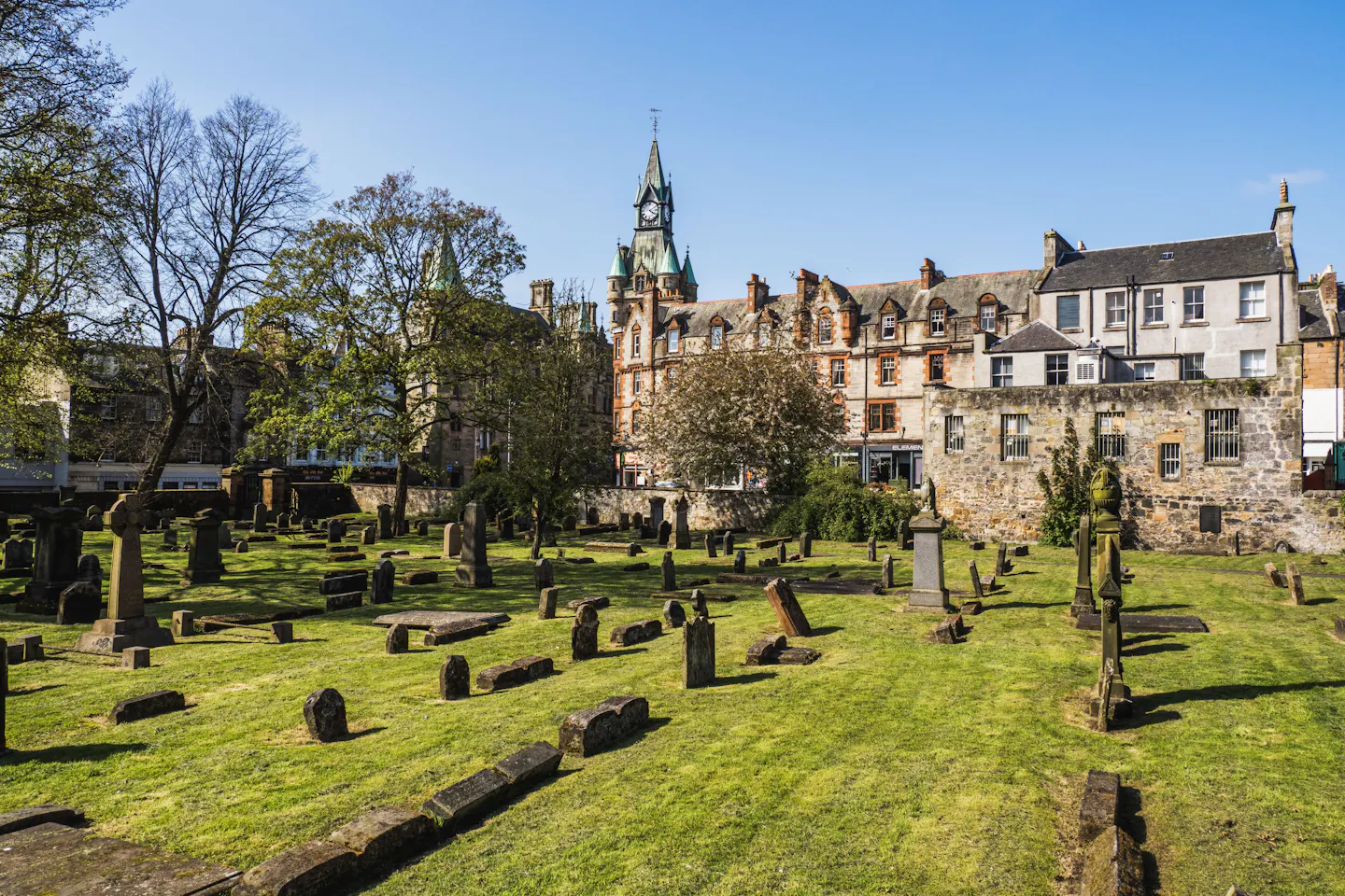 Greyfriars Kirkyard: weathered upright and recumbent gravestones across mown grass, ringed by Old Town tenements and a green-domed clock tower