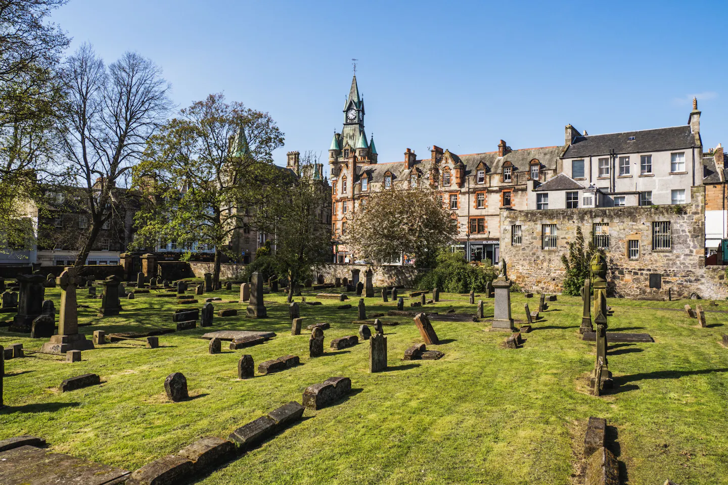 Greyfriars Kirkyard: weathered upright and recumbent gravestones across mown grass, ringed by Old Town tenements and a green-domed clock tower