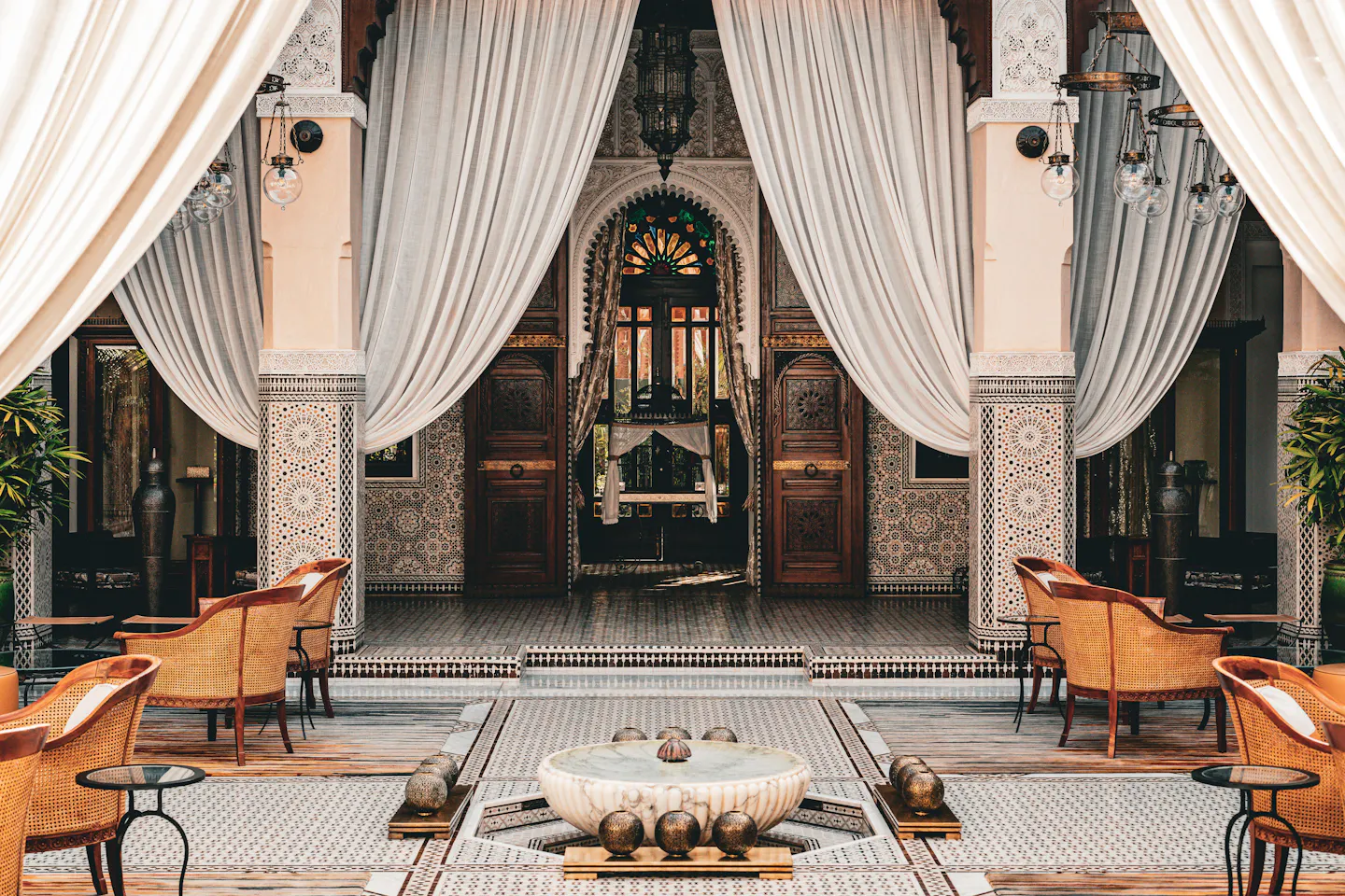 A Marrakech riad courtyard with zellige tilework, carved arches and white drapes around a marble fountain