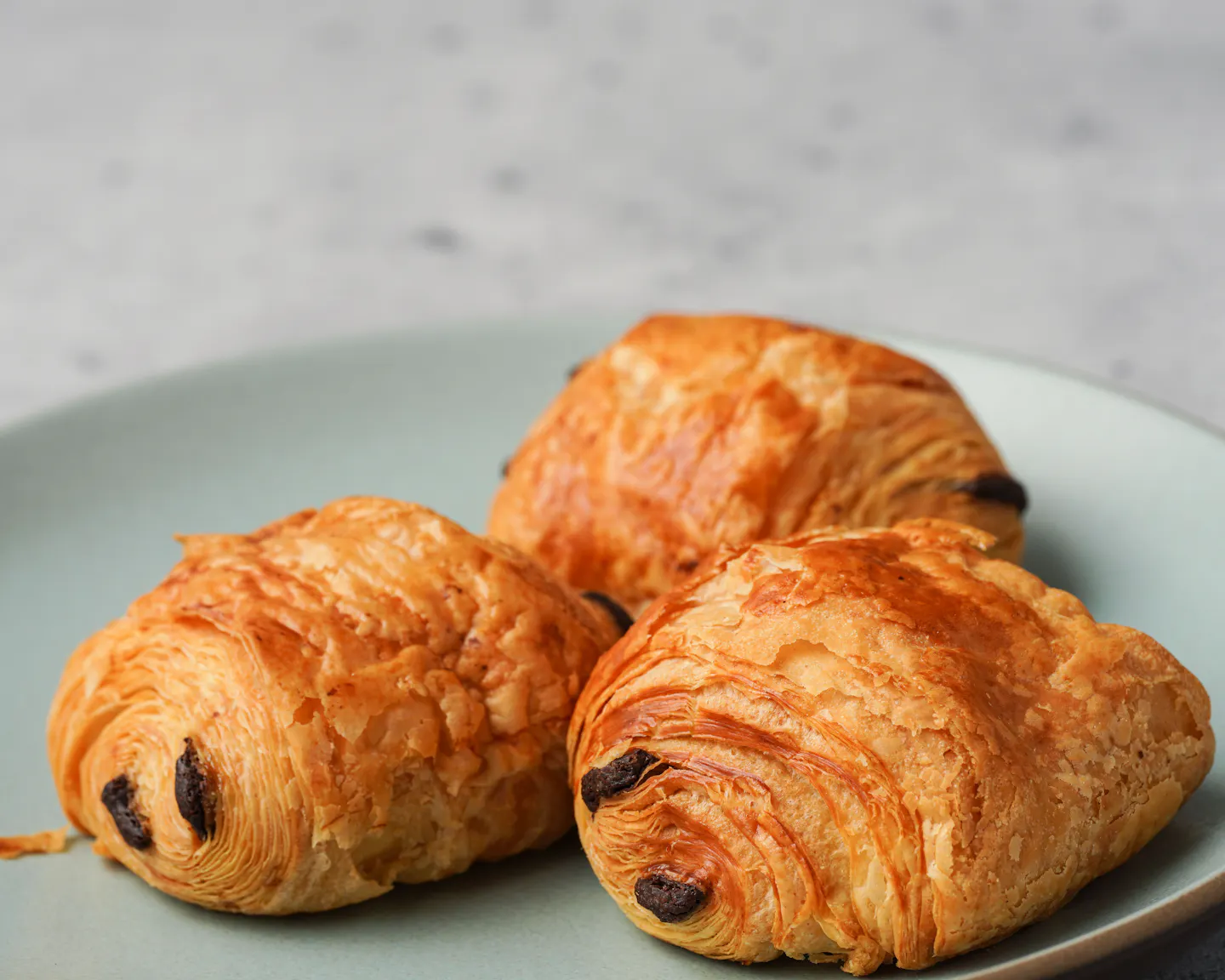 Three freshly baked pain au chocolat with visible flaky layers and dark chocolate batons, on a pale sage ceramic plate