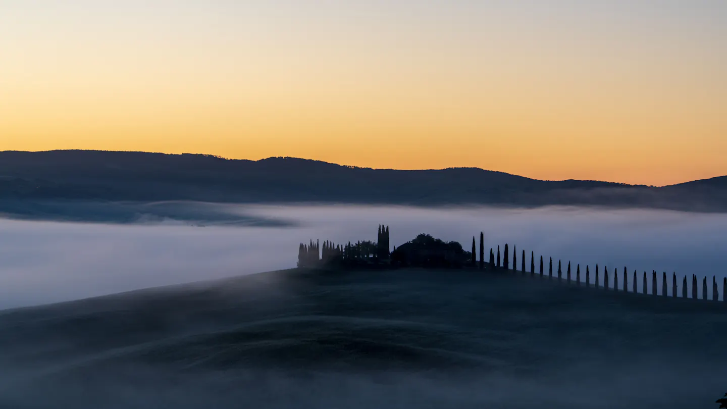 Misty rolling hills of Tuscany's Val d'Orcia at dawn, with a hilltop farmhouse and a line of cypress trees