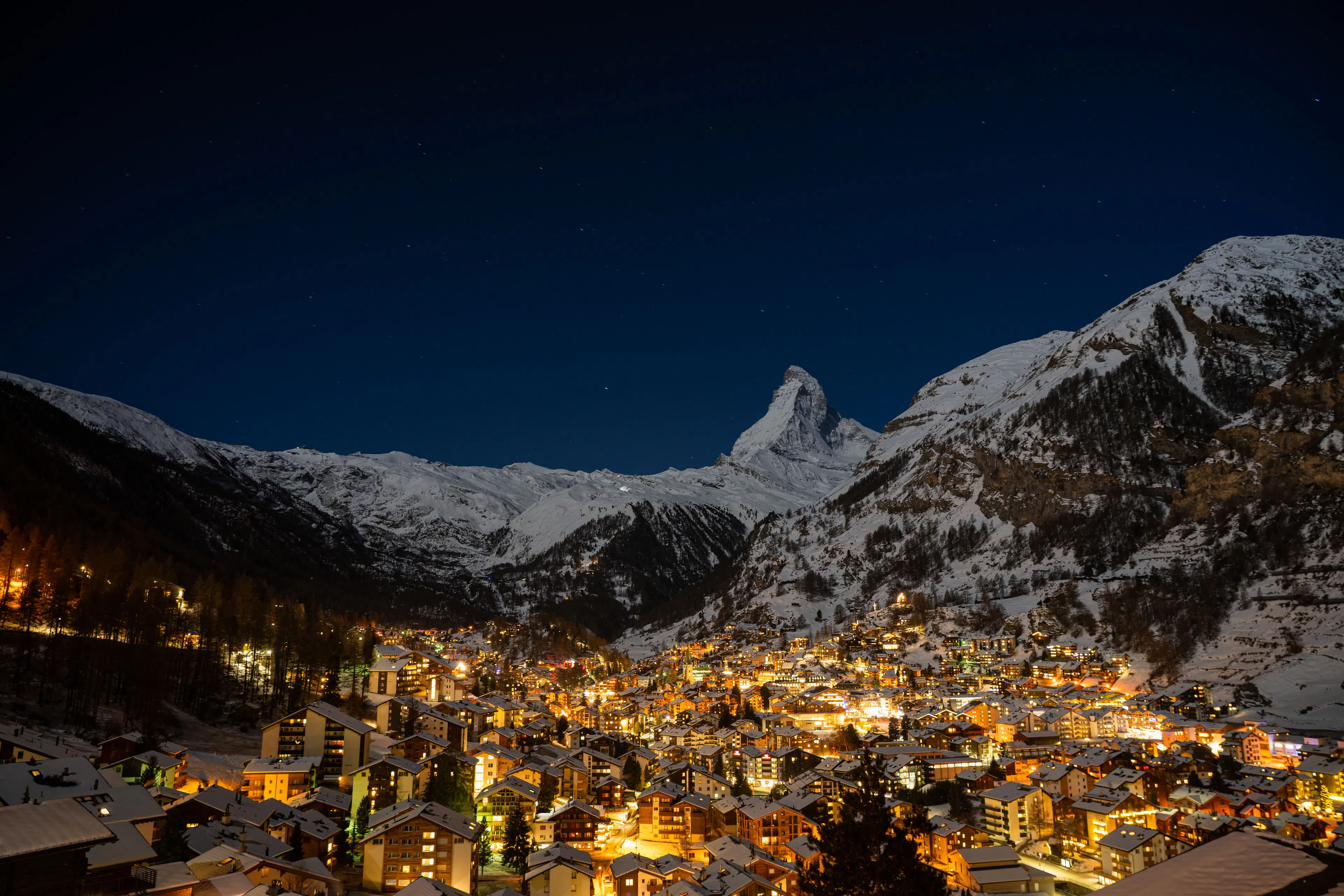 Zermatt village glowing at night beneath the floodlit Matterhorn, warm chalet windows and street lamps under a star-filled deep-blue sky