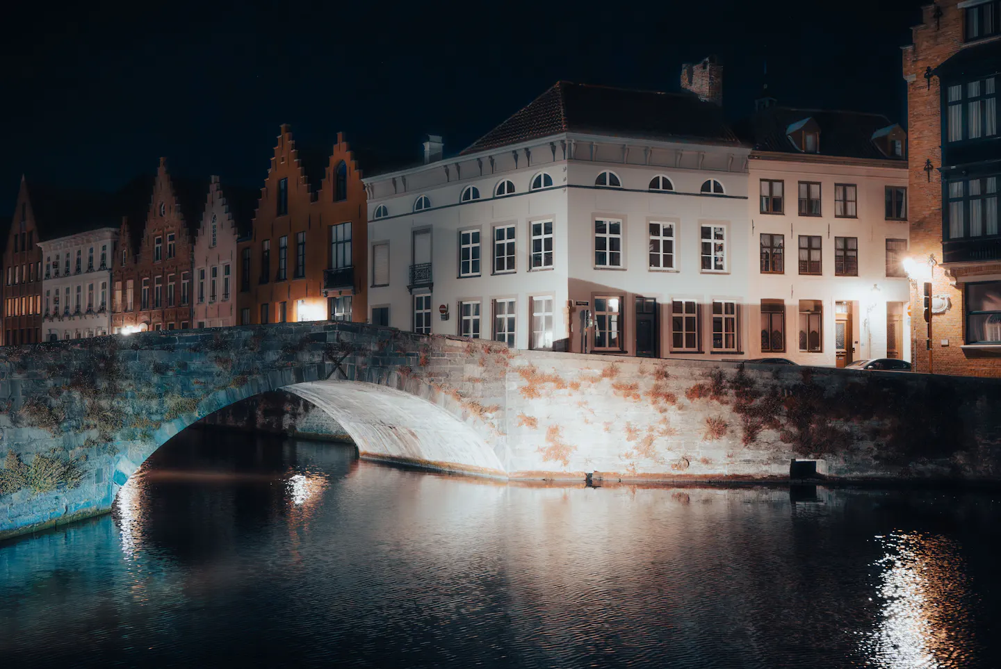 A stone humpback bridge and a row of stepped-gable Bruges houses lit up at night, their warm windows and lamps rippling in the dark canal below