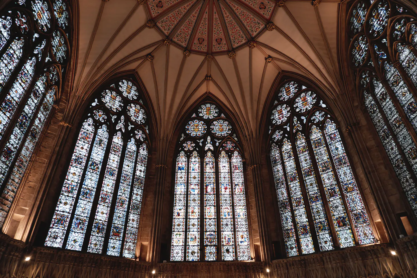 Soaring stained-glass windows and a painted vaulted ceiling inside York Minster, five tall traceried lights filled with medieval glass above a carved stone arcade