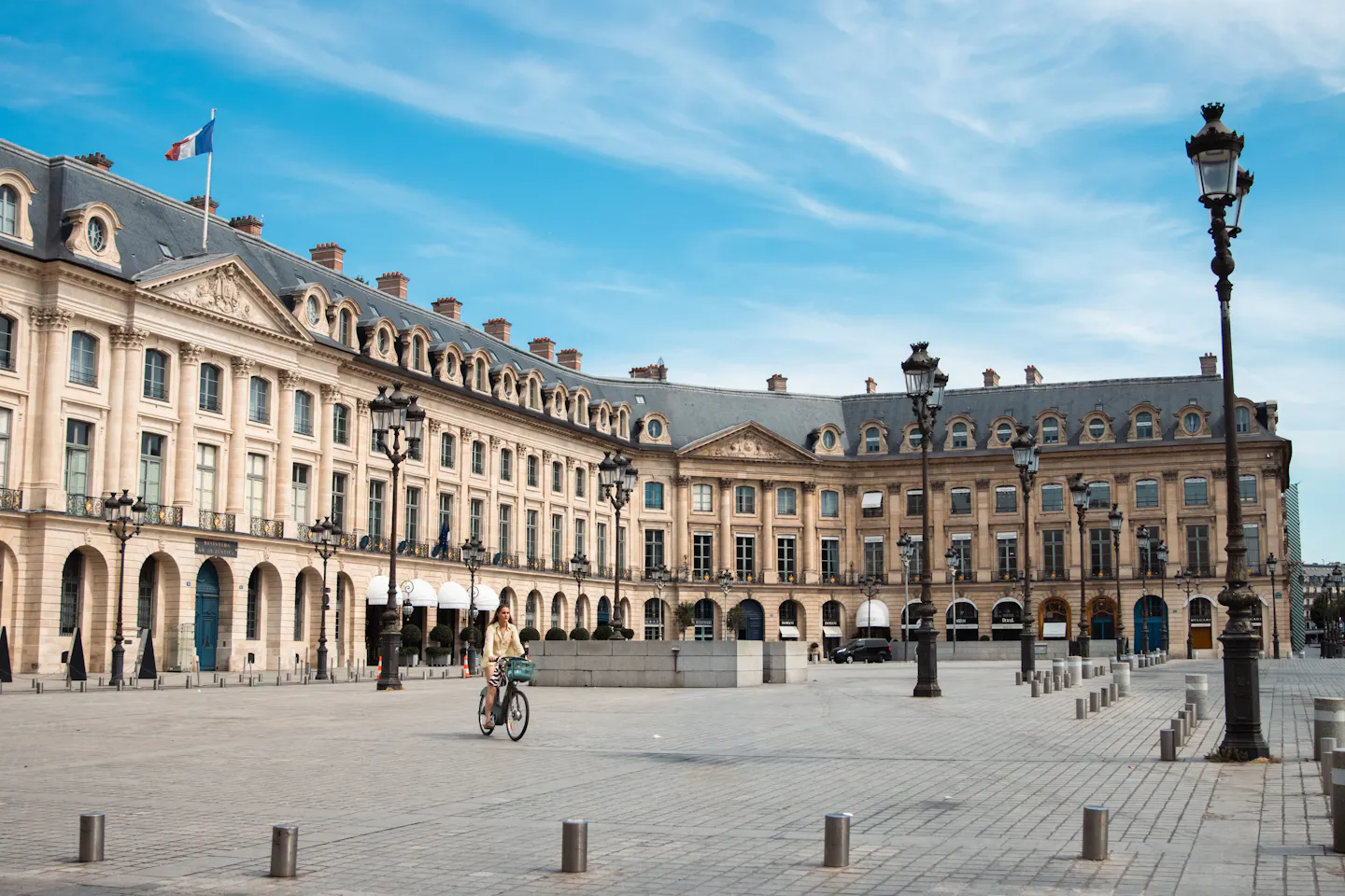 A cyclist crossing a broad, empty Paris square ringed by arcaded stone facades and ornate lamp posts under a blue sky
