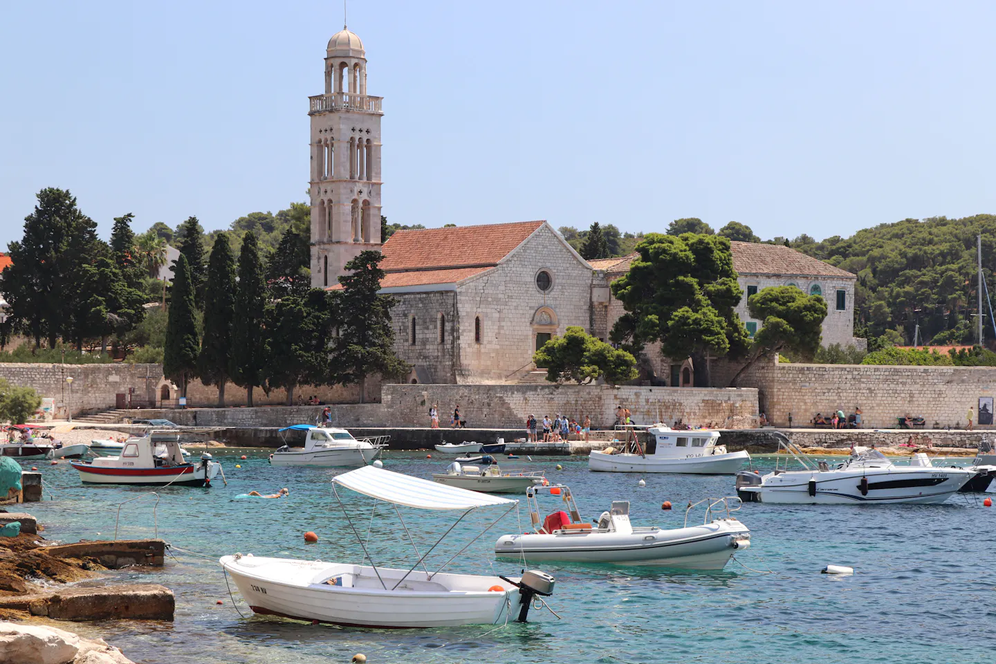The stone bell tower of the Franciscan monastery in Hvar above cypresses and small boats moored in clear turquoise water