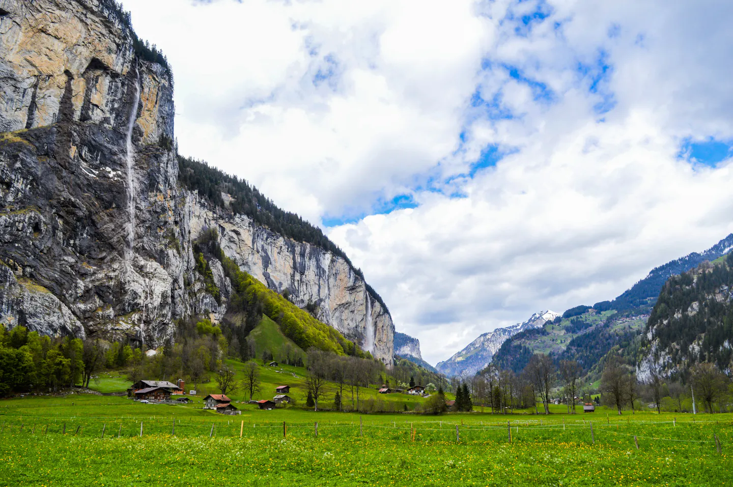 The Lauterbrunnen valley floor: a flower-dotted green meadow and wooden chalets beneath sheer limestone cliffs with waterfalls dropping down the rock, snow peaks closing the valley under blue sky and cumulus