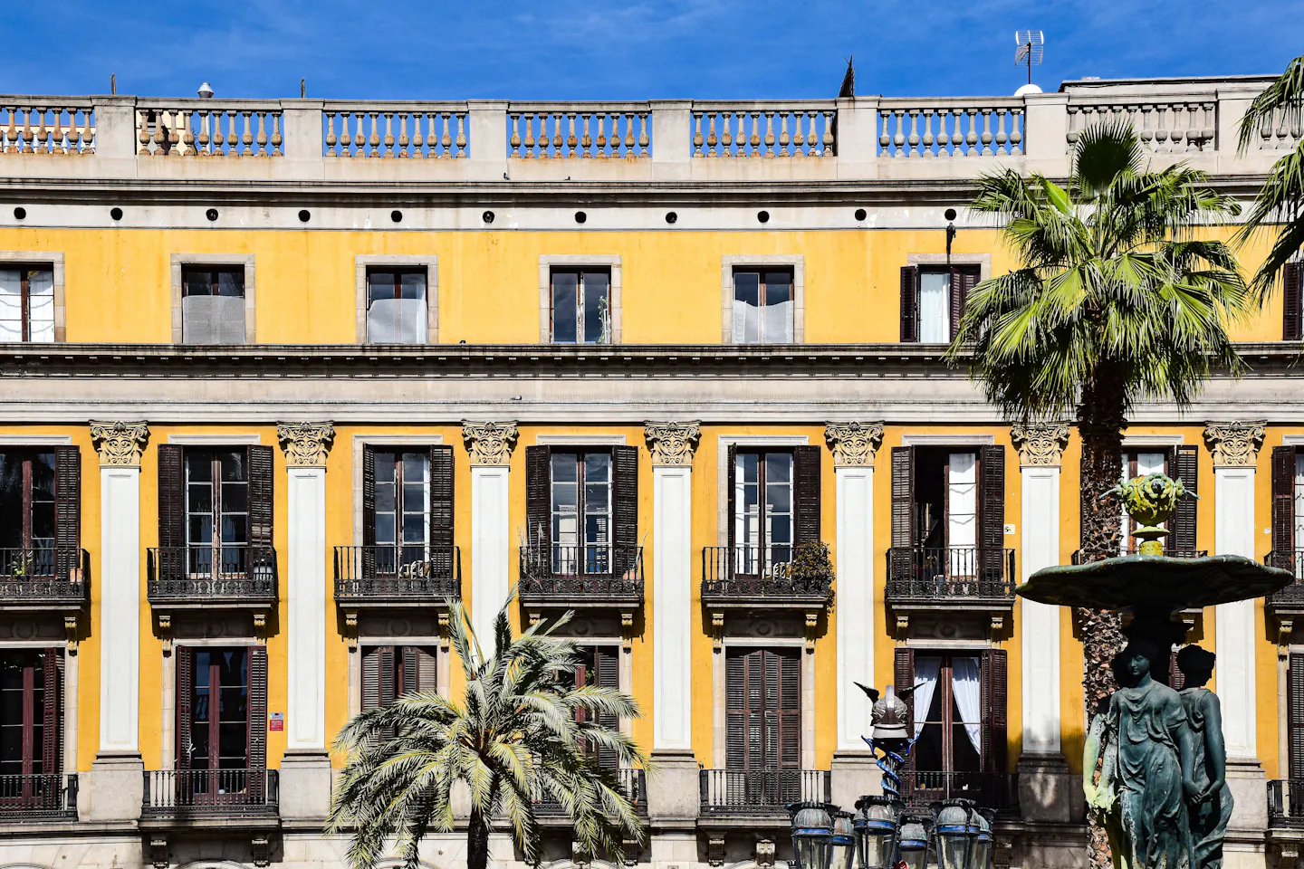 Palm trees and an ornate lamppost in front of the yellow arcaded facade of Placa Reial in Barcelona