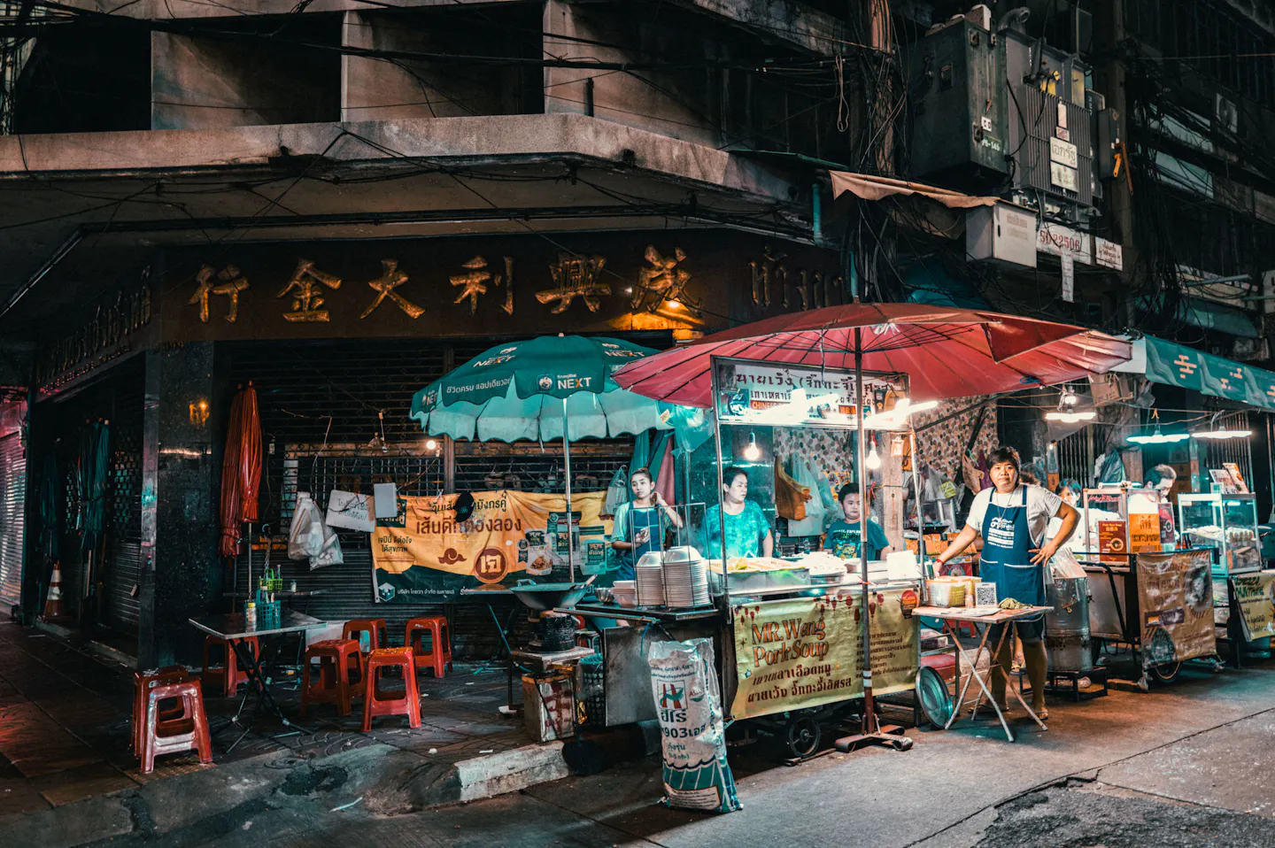 Street food stalls with red and green umbrellas, stools and vendors on a Bangkok Chinatown corner at night