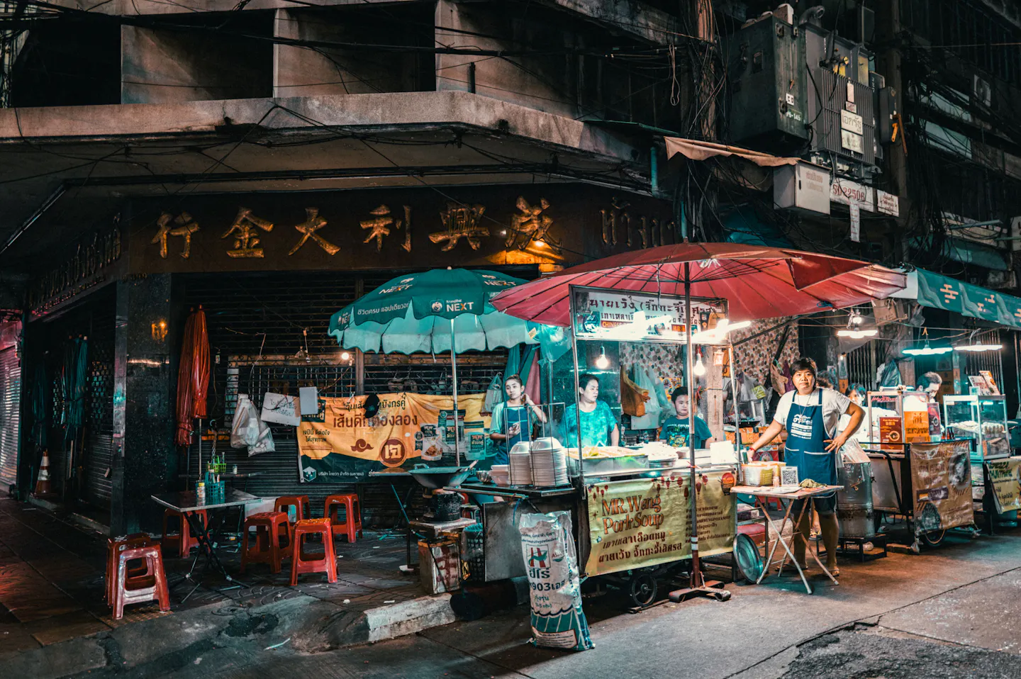 Street food stalls with red and green umbrellas, stools and vendors on a Bangkok Chinatown corner at night