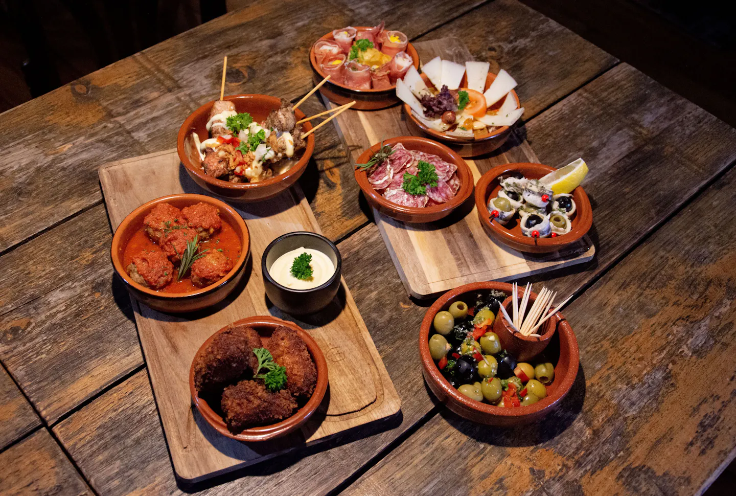 Terracotta dishes of croquetas, meatballs, olives, cured ham and cheese spread across a rustic wooden table