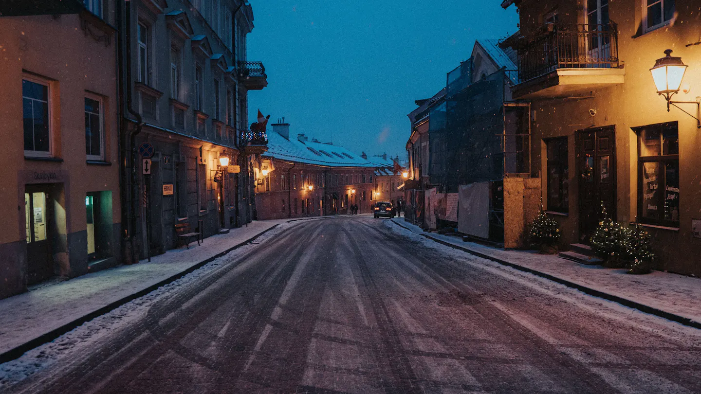 Snow covering a lamplit Old Town street in Vilnius at dusk, with small lit Christmas trees beside a doorway