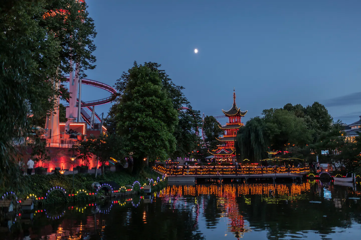 The illuminated pagoda, roller coaster and strings of lights of Tivoli Gardens reflected in the lake at dusk in Copenhagen