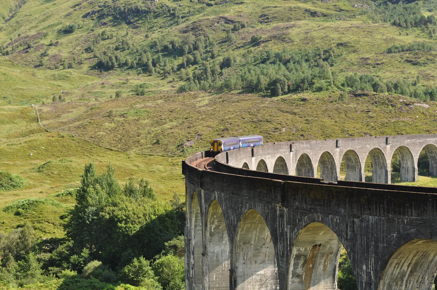 A train crossing the curved stone Glenfinnan Viaduct through green Highland hills