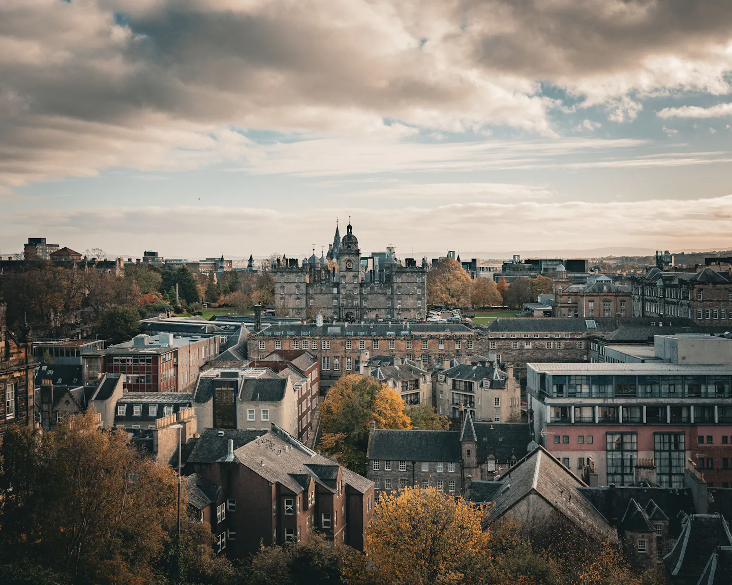 Edinburgh’s stone rooftops, chimneys and spires among orange and gold autumn trees under a wide cloudy sky