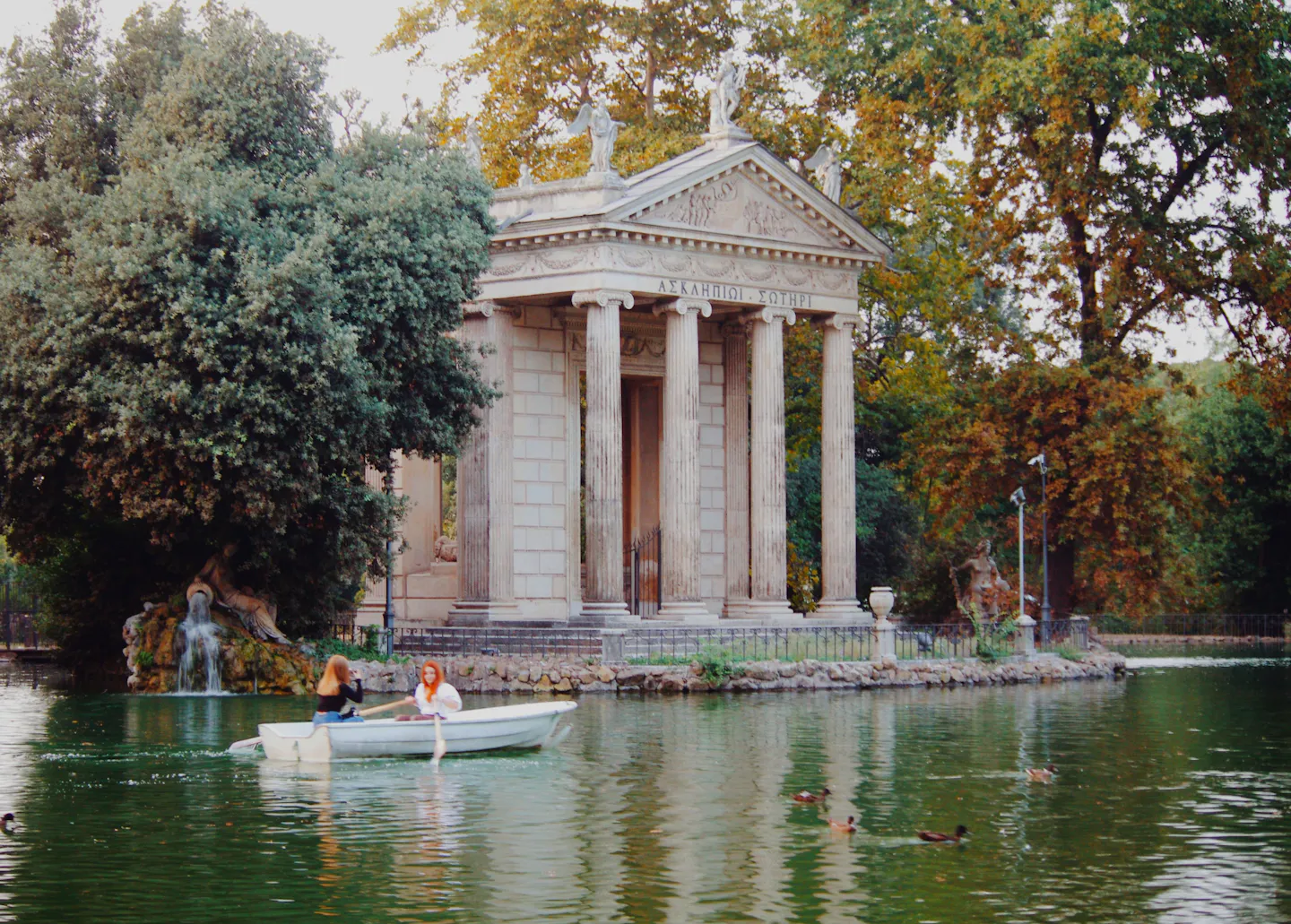 The Temple of Aesculapius on the lake in the Villa Borghese gardens, Rome, with a rowing boat on the green water and tall trees around it