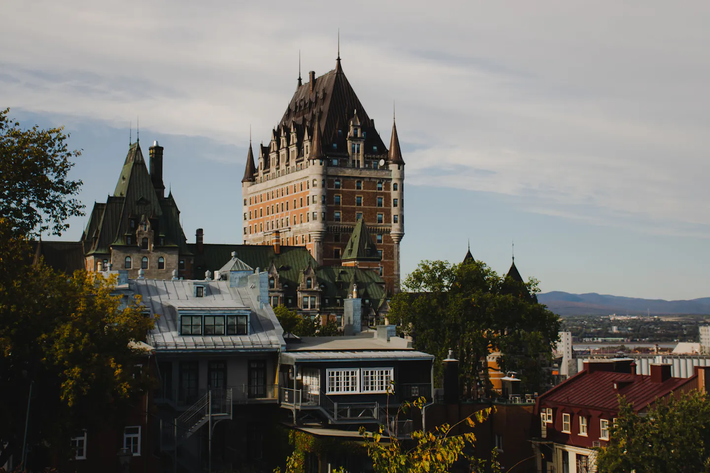 Château Frontenac's copper turrets and brick facade rising above the steep grey roofs and dormer windows of Old Québec in warm afternoon light, hills across the St Lawrence in the distance