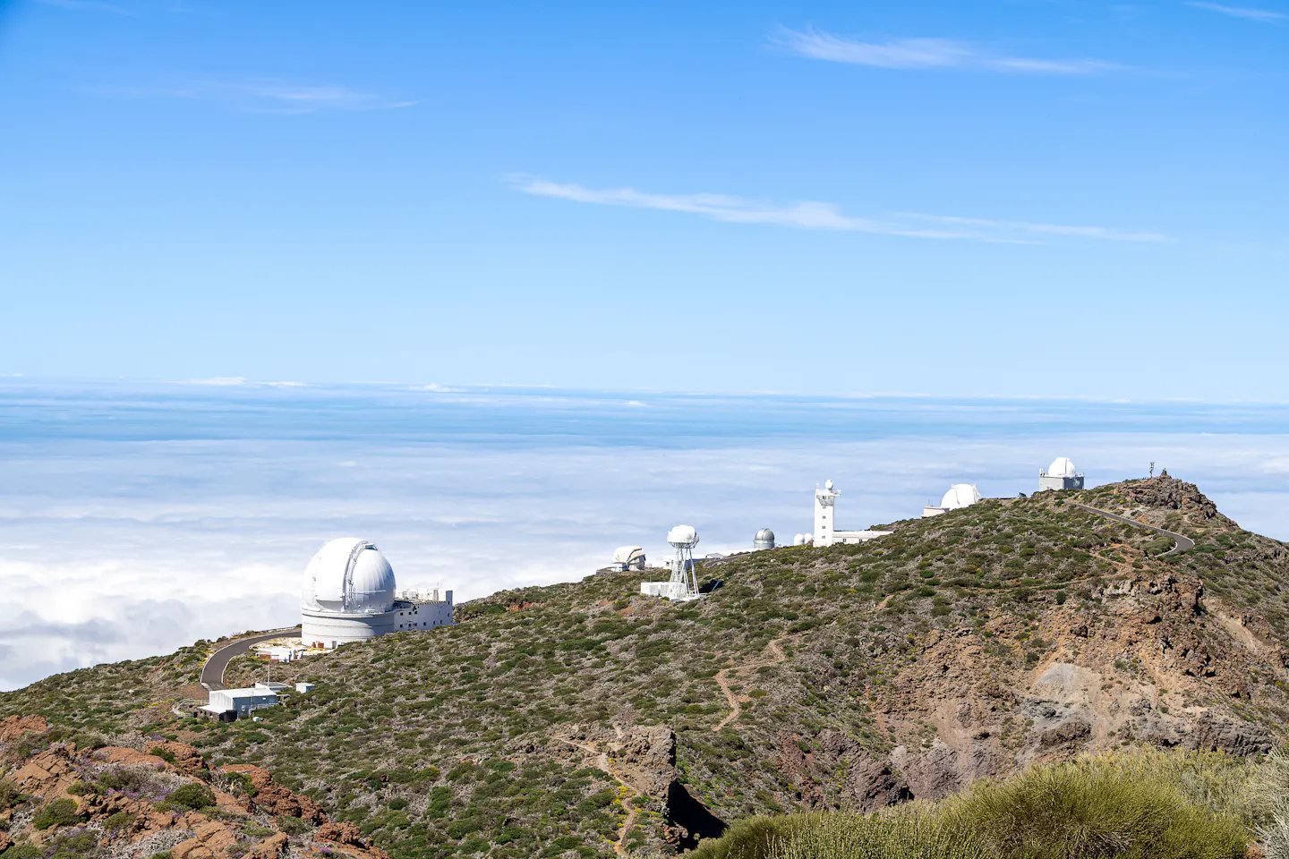 White telescope domes strung along the Roque de los Muchachos ridge on La Palma, standing above a sea of cloud under a deep blue sky