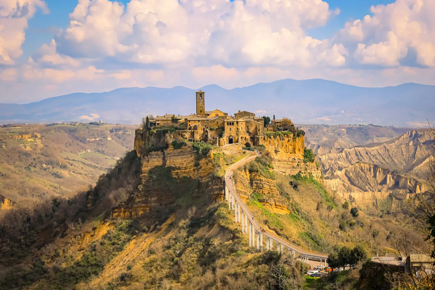 A stone hilltop village on a sheer tufa outcrop in the Lazio countryside, reached by a long footbridge, with eroded ravines and blue hills behind under a big cloudy sky