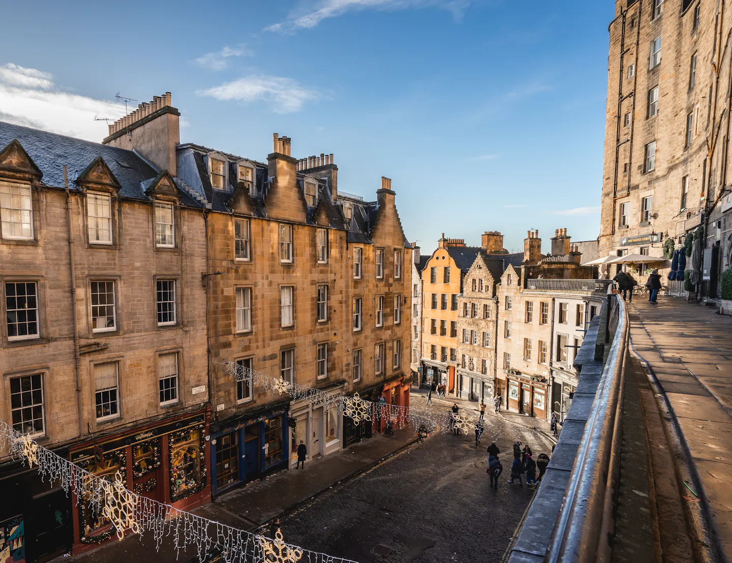 Curved terrace of honey-coloured Old Town tenements above Edinburgh's West Bow, strung with snowflake lights and lit by low winter sun, the cobbled street curving away below