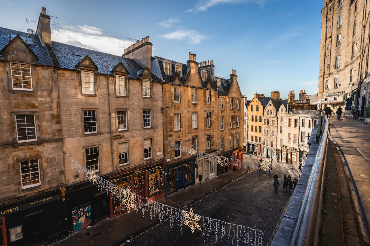 Curved terrace of honey-coloured Old Town tenements above Edinburgh's West Bow, strung with snowflake lights and lit by low winter sun, the cobbled street curving away below