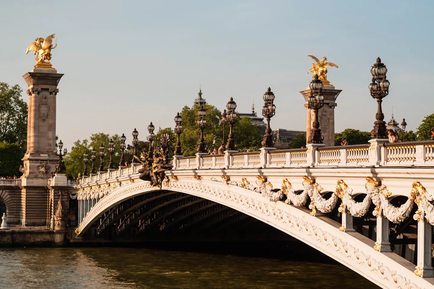 The gilded swags and white stone arch of Pont Alexandre III in warm evening light, its ornate lamp posts and two golden winged statues on their columns above the Seine