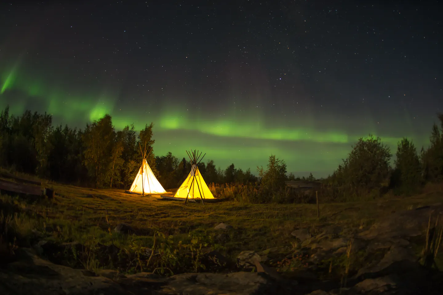 Two lamp-lit tipis at an aurora camp outside Yellowknife, Canada, under a green band of northern lights
