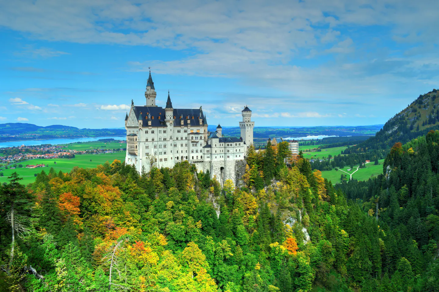Neuschwanstein Castle above a forest turning orange and gold, with Bavarian lakes and farmland beyond