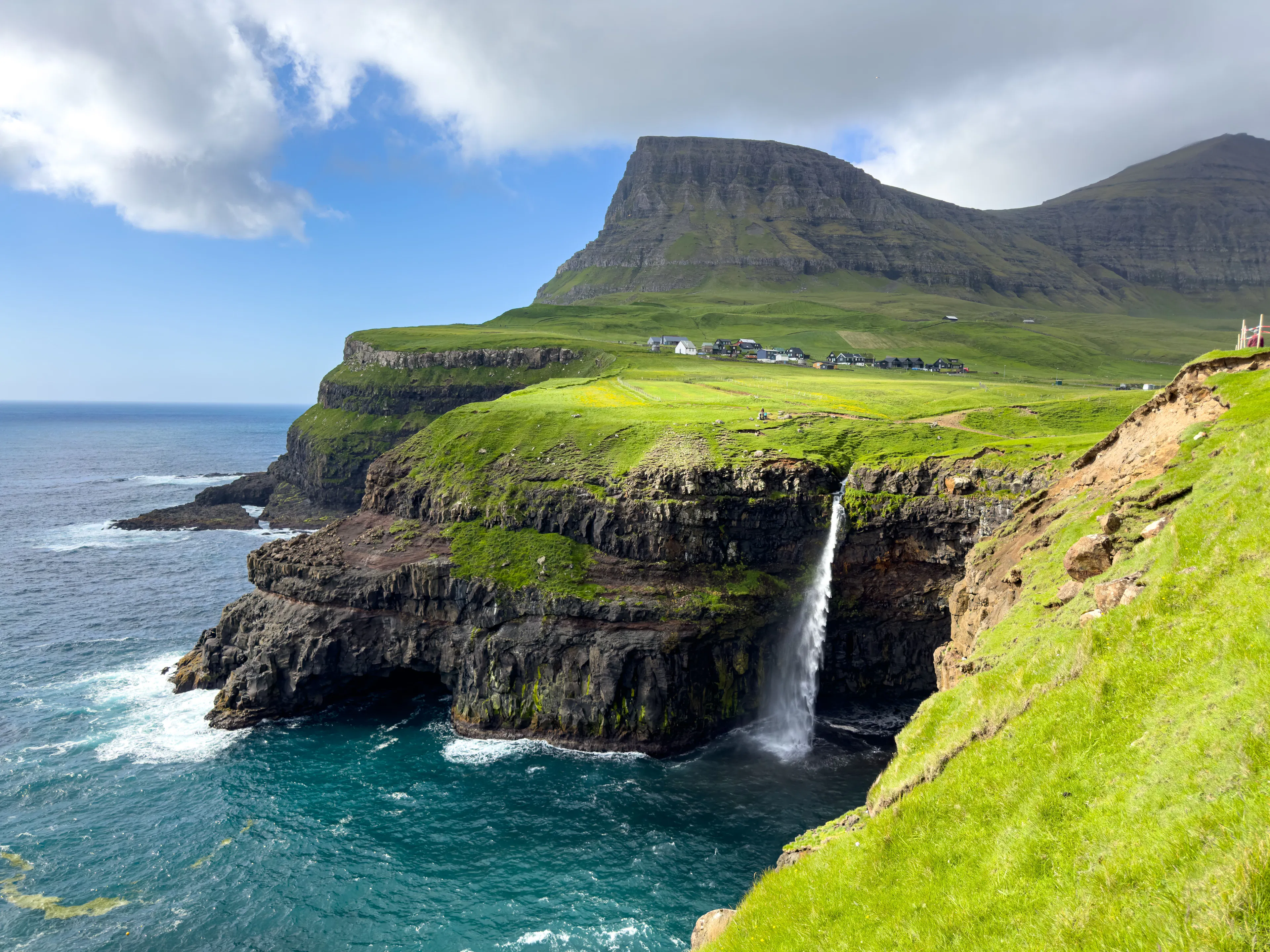 Múlafossur waterfall dropping from a green clifftop into the turquoise Atlantic beside the village of Gásadalur in the Faroe Islands