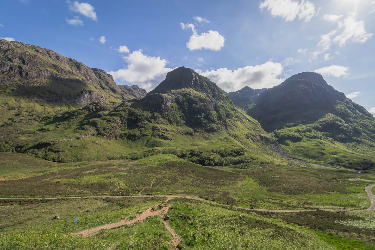 The Three Sisters ridges of Glencoe rising green and sunlit above a moorland path in the Scottish Highlands
