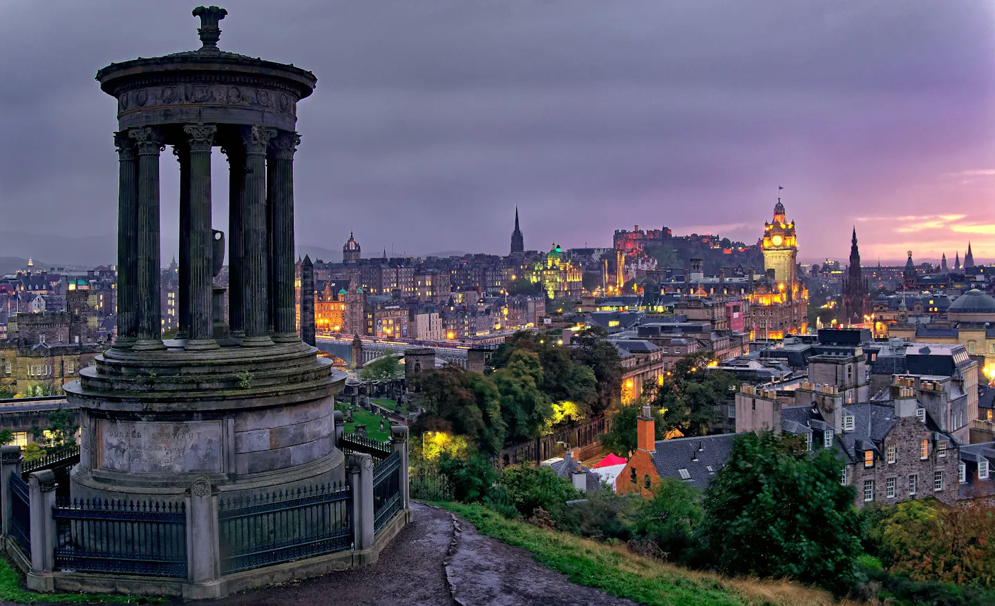 Calton Hill at blue hour: the columned Dugald Stewart Monument in the foreground with the Old Town skyline, the castle and the floodlit Balmoral clock tower glowing beneath a violet sky