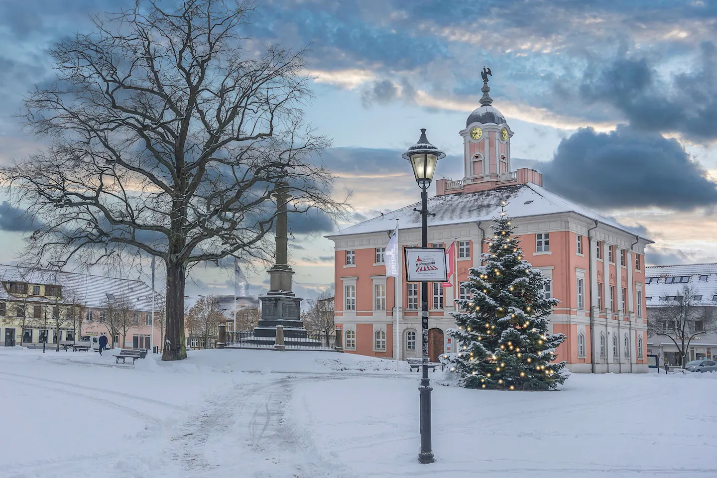 A decorated Christmas tree on a snow-covered square beside a pink baroque town hall at dusk