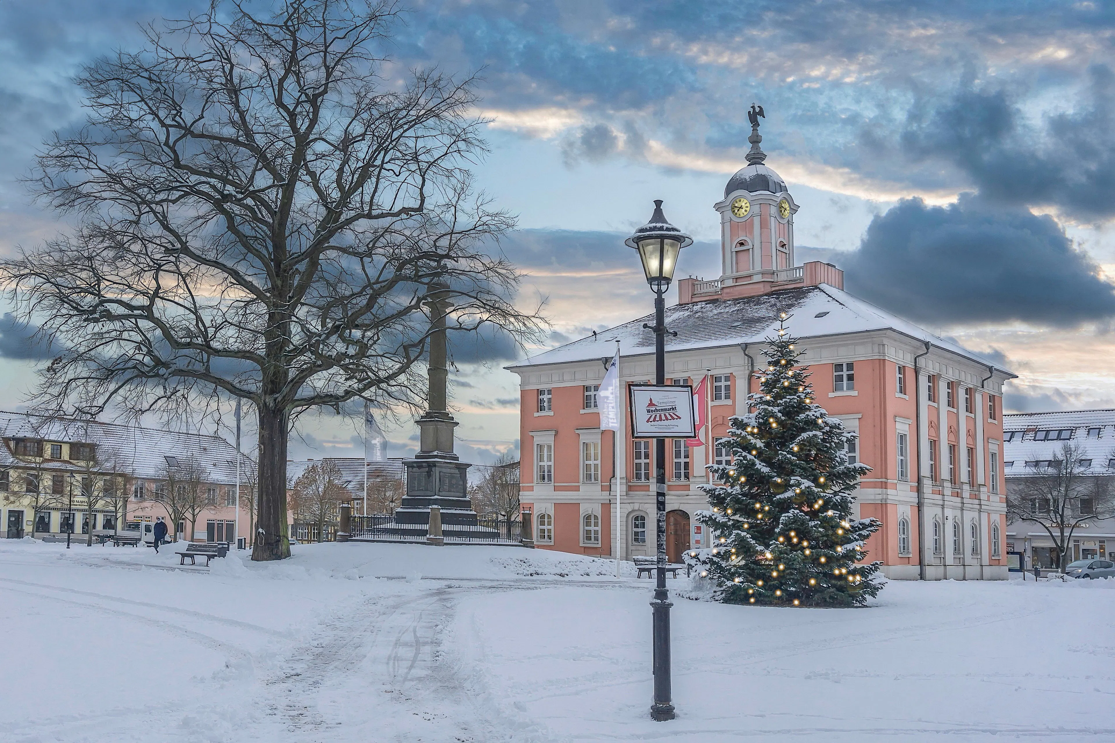 A decorated Christmas tree on a snow-covered square beside a pink baroque town hall at dusk