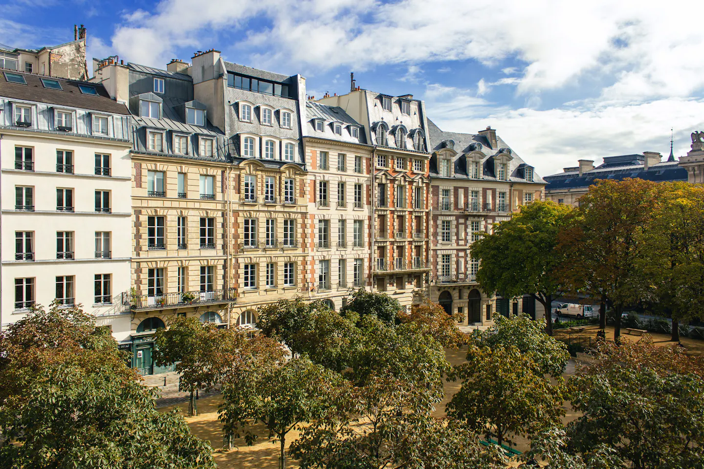 An elevated view over a quiet tree-lined Paris square, stone and brick townhouses with steep mansard roofs on three sides under a blue sky with broken cloud