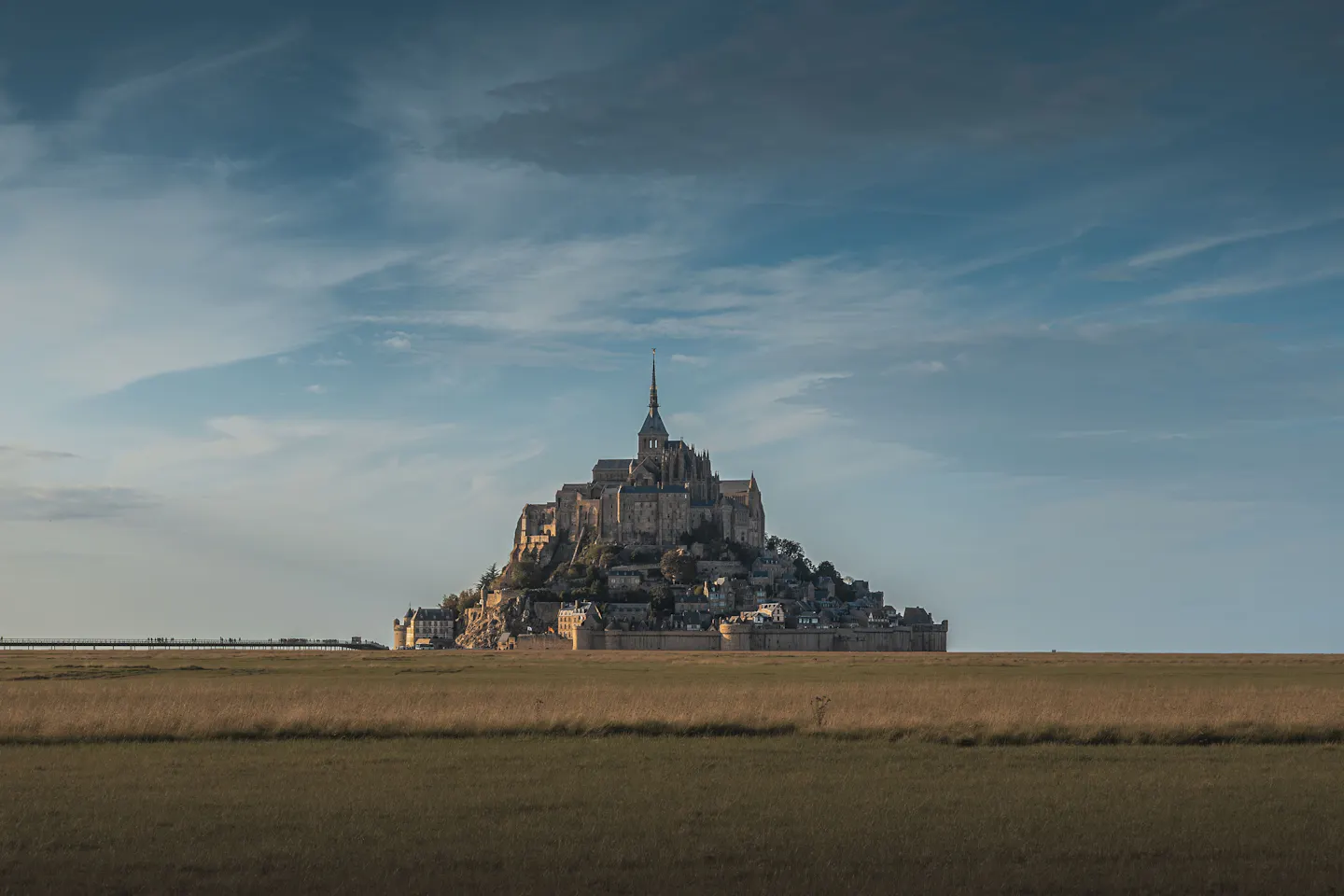 Mont-Saint-Michel rising from flat salt marsh at dusk, its abbey spire and ramparts catching the last warm light under a wide sky of wispy cloud
