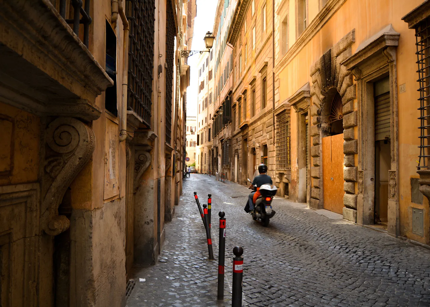 A rider on a scooter turning down a narrow cobbled Roman lane between tall ochre palazzi with carved stone doorcases and shuttered windows, in warm afternoon light