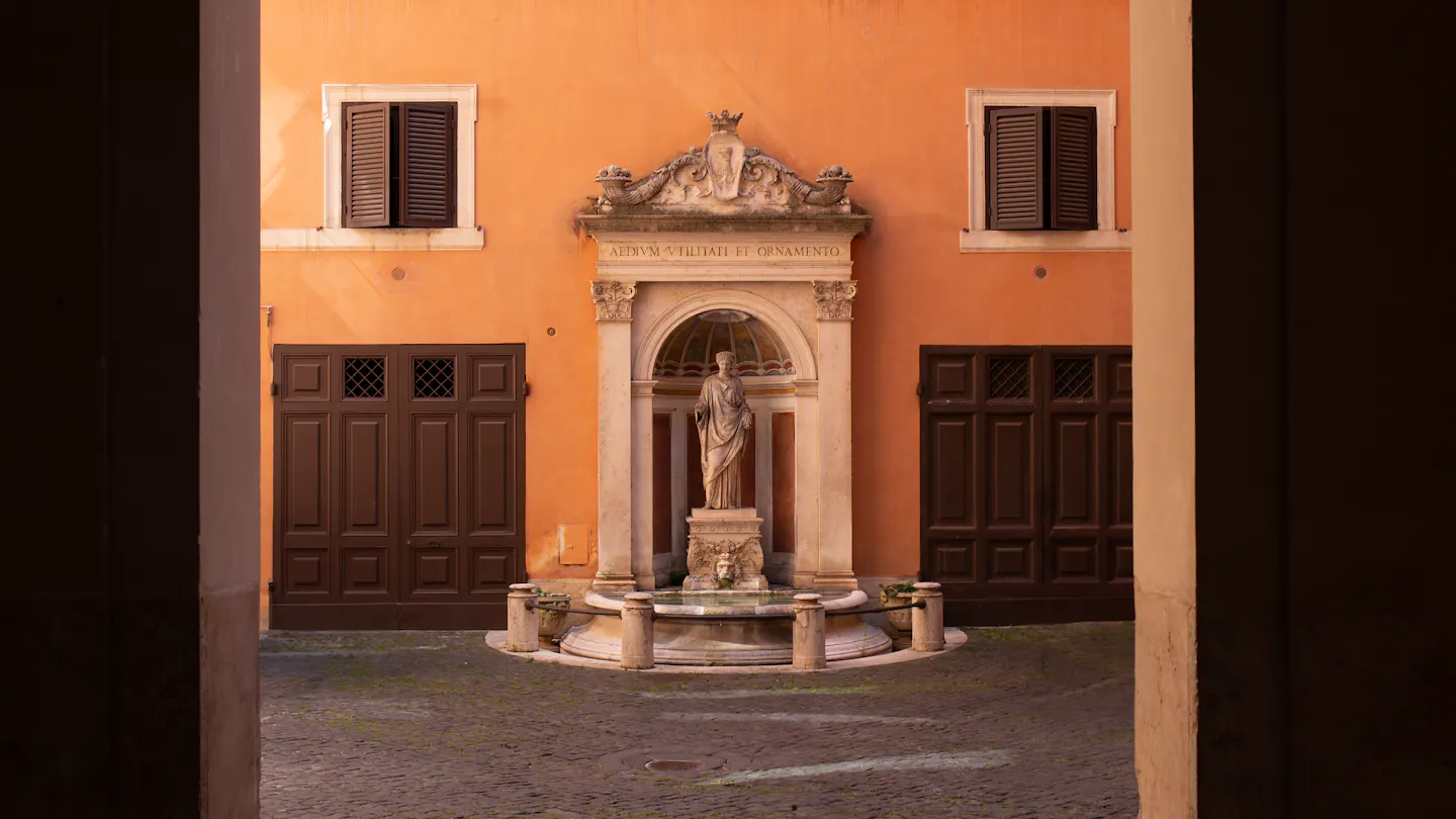 An ornate stone niche fountain with a robed statue set into a burnt-orange palazzo wall, seen through a dark archway across a cobbled courtyard