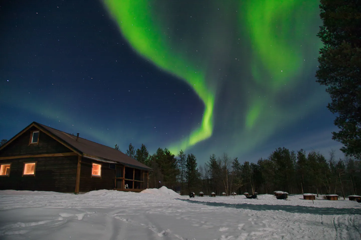 A wooden cabin with lit windows under a green aurora in snowy Swedish Lapland