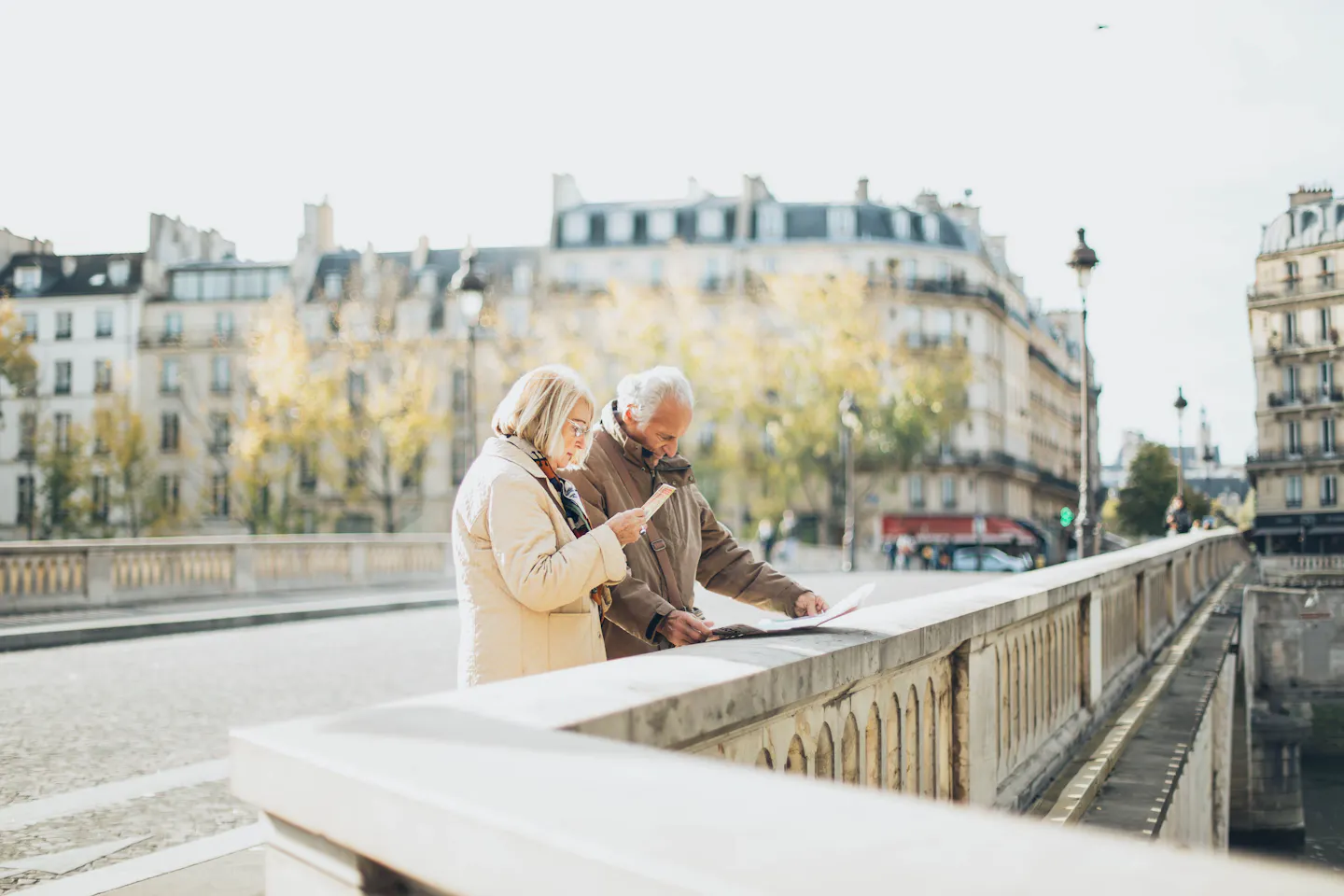A couple checking a paper map and a smartphone on a bridge in Paris, with Haussmann-style buildings behind